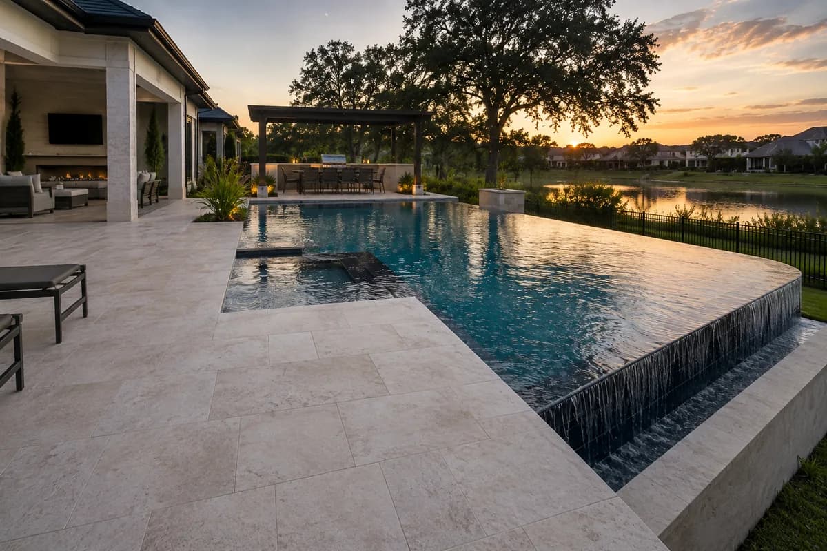 Infinity-edge pool spilling toward a lake at sunset with covered patio and pergola